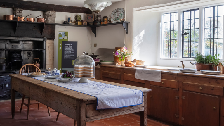 A wide shot of the kitchen at Avebury Manor, with a table laid with cups, teapot and large cake, with flowers and brass pots and pans arranged around a large stove